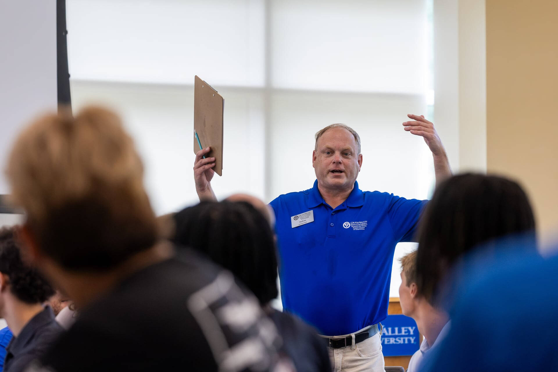 Tim Born, in blue GVSU polo, gestures with arms raised in front of students who are seated